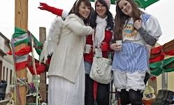 Drei Frauen in Winterkleidung und Kostümen stehen in einer festlichen Umgebung im Freien. Eine Frau hat Flügel, eine hält einen Becher, und sie sind von bunten Fahnen umgeben.