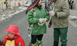 Eine Frau in einer grünen Jacke und ein Mann in grüner Hose und einer roten Mütze stehen auf der Straße, mit einem Kind in Rot vor ihnen.