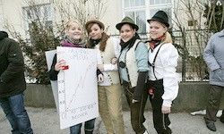 Vier Frauen in Vintage-Kleidung stehen zusammen draußen vor einem Gebäude und Zaun und halten ein Stück Papier mit einer Zeichnung in der Hand.