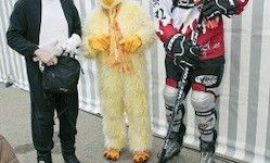 Drei Personen in Kostümen stehen vor einem gestreiften Hintergrund. Einer trägt einen schwarzen Anzug, ein anderer ein Hühnerkostüm und der dritte die Ausrüstung eines Eishockeyspielers.