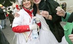 Zwei Frauen in Kostümen, eine in Weiß und Rot mit rotem Schal, und die andere in Schwarz, posieren für die Kamera bei einer festlichen Veranstaltung.