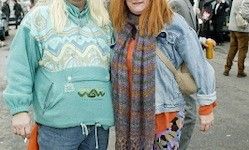 Zwei Frauen stehen in einem Außenbereich, vermutlich auf einer Straße. Die Frau auf der linken Seite raucht eine Zigarette und trägt eine grüne Jacke. Die andere Frau trägt eine Jeansjacke, einen Schal und eine Hose.