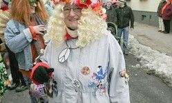 Eine Frau mit blonder Lockenperücke und Brille steht auf einer Straße mit bunten Dekorationen. Hinter ihr sind andere in Kostümen gekleidet. Schnee liegt auf dem Boden.