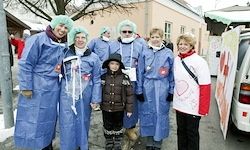 Eine Gruppe von Menschen in blauen medizinischen Kitteln und grünen Haarnetzen steht zusammen vor einem Gebäude. Ein Kind in einer braunen Jacke und Mütze steht in der Mitte.