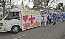 Ein weißer Lieferwagen mit rotem Kreuz und 'Drs. auf dem Weg' fährt auf der Straße, mit medizinischen Fachkräften in blauen Uniformen, die hinter ihm gehen.