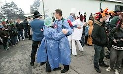 Eine Gruppe von Menschen in Kostümen versammelt sich im Freien, einige tanzen in der Mitte. Einige tragen blaue Regenmäntel, Hüte und Stiefel. Dahinter beobachtet eine Menge. Bäume und ein Gebäude sind im Hintergrund.