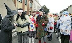 Menschen in bunten Kostümen und Hüten stehen auf der Straße, möglicherweise bei einem Festival. Einige tragen Stiefel und Handschuhe. Gebäude säumen die Straße.