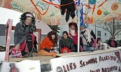 Eine Gruppe von Menschen in Kostümen posiert für ein Foto auf einer Bühne, mit farbenfrohen Dekorationen und einem orangefarbenen Banner mit der Aufschrift Alles Sehnsehe Alles Mehr.