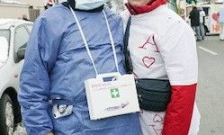Zwei Personen in medizinischen Kleidungsstücken stehen im Freien. Eine trägt eine blaue Robe und eine Maske und hält eine Erste-Hilfe-Box. Die andere Person ist in weiß und rot gekleidet mit einem Herzsymbol. Ein Auto und Personen sind im Hintergrund.