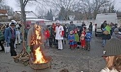 Eine Menschenmenge versammelt sich um einen brennenden Feuerkorb, mit einer Person in einem weißen Outfit in der Mitte. Kinder in bunten Kostümen und Erwachsene sind anwesend, einige halten Gegenstände. Ein weißer Lieferwagen ist geparkt.