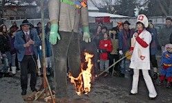 Eine große Figur, die einen grünen Handschuh trägt, steht in Flammen, während eine Frau in einem weißen Mantel ein Mikrofon hält. Eine Menschenmenge in festlicher Kleidung beobachtet.