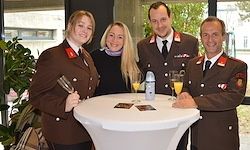 Vier Personen in Militäruniformen posieren für ein Foto. Zwei Frauen und zwei Männer stehen hinter einem runden Tisch. Zwei Champagnergläser stehen auf dem Tisch. Ein Topf mit Pflanze im Hintergrund.