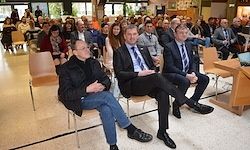 Drei Männer in Anzügen sitzen auf Stühlen vor einem sitzenden Publikum. Zwei Männer haben die Beine verschränkt, der dritte hat die Arme verschränkt.