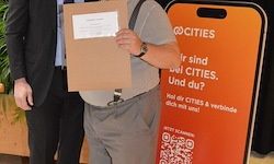 Ein Mann in grauen Shorts hält eine braune Mappe mit einem weißen Papier vor einem orangefarbenen Schild, das '@CITIES' und 'Bist du auch eine Stadt?' mit einem QR-Code zeigt.