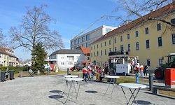 Ein gepflasterter Bereich mit mehreren Tischen und Stühlen, umgeben von Bäumen und Gebäuden. Menschen sind um ein Fahrzeug versammelt, möglicherweise eine Straßenbahn. Der Himmel ist klar und blau.