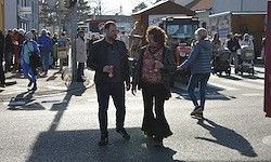 Ein Mann und eine Frau gehen gemeinsam auf einer belebten Straße. Viele Menschen schlendern und shoppen um sie herum, mit Straßenverkäufern und einigen Fahrzeugen im Hintergrund.