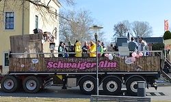 Ein dekorierter Lastwagen mit einer Menschenmenge auf dem Anhänger, beschriftet mit 'Schwaiger Alm', steht auf der Straße geparkt.