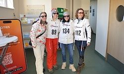 Vier Frauen in Winterkleidung stehen in einem Flur. Sie tragen die Nummern 89, 134 und 112 auf ihren Jacken. Sie lächeln alle und posieren für ein Foto.