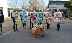 Eine Gruppe von Menschen in Kostümen schiebt einen Wagen mit bunten Luftballons. Sie stehen auf einem Parkplatz mit Gebäuden und Bäumen im Hintergrund.