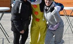 Zwei Frauen posieren für ein Foto mit einer Person in einem Clownkostüm, die auf einem gepflasterten Bereich mit runden Tischen steht.