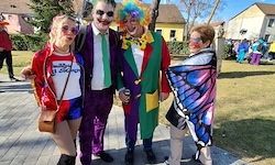 Vier Menschen in farbenfrohen Kostümen posieren für ein Foto in einem Park mit Häusern und Bäumen im Hintergrund.