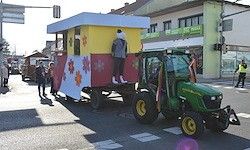 Ein grüner Traktor zieht eine bunte Deko auf der Straße, auf der eine Person steht. Ein Fußgänger geht in der Nähe vorbei, und Gebäude sind im Hintergrund.