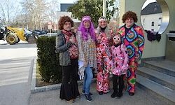 Eine Gruppe von Menschen in bunten Kostümen posiert vor einem Gebäude mit einem runden Spiegel und einem gelben Baufahrzeug, das auf der Straße geparkt ist.