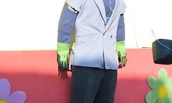Eine Person in einem weißen Mantel und grünen Handschuhen steht auf einer roten Oberfläche mit dekorativen Blumen. Der Hintergrund ist weiß.