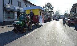 Ein dekorierter Traktor fährt mit einer Person darauf sitzend die Straße hinunter. Es gibt Leute, die auf dem Bürgersteig gehen, und einige stehen in der Nähe geparkter Autos. Links von der Straße befinden sich Gebäude.