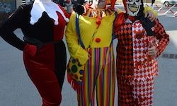 Drei Menschen in Kostümen stehen auf einer Straße zusammen. Einer trägt ein Joker-Kostüm, einer ein Clown-Kostüm und der andere ein Harlekin-Kostüm. Sie scheinen für ein Foto zu posieren.