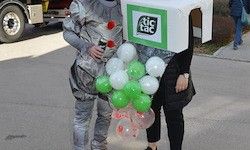 Eine Person in einem silbernen Anzug, der einen Strauß grüner und weißer Luftballons hält, und eine Schachtel mit den Worten 'tic tac' darauf.