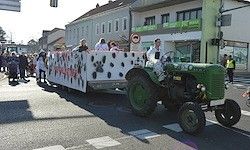 Ein grüner Traktor mit einem geschmückten Anhänger, der Menschen transportiert, fährt durch eine Stadtstraße mit Gebäuden und Zuschauern.