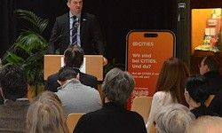 Ein Mann in Anzug und Krawatte steht an einem Rednerpult und spricht zu einem sitzenden Publikum. Hinter ihm sind eine Pflanze und ein rotes Schild mit dem Text 'Wir finden Städte' zu sehen. Das Publikum besteht aus Männern und Frauen, von denen einige aufmerksam zuhören.