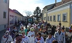 Ein Umzug von Menschen in Kostümen geht die Straße entlang. Viele tragen Masken. Es gibt Gebäude auf beiden Seiten der Straße.