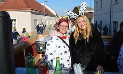 Zwei Frauen stehen bei einem Tisch auf einem Straßenfest. Eine Frau trägt ein Dalmatiner-Kostüm und die andere einen Pelzmantel. Flaschen, Gläser und ein schwarzer Eimer sind auf dem Tisch. Im Hintergrund sind Gebäude und Menschen zu sehen.