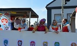 Eine Gruppe von Menschen in passenden Uniformen steht hinter einer weißen Barriere, die mit Zeichentrickfiguren verziert ist. Sie lächeln und posieren für ein Foto. Hinter ihnen sind Häuser und eine Ampel.