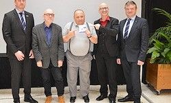 Sechs Männer in Anzügen und Krawatten stehen in einem Konferenzraum. Ein Mann hält eine Karte.