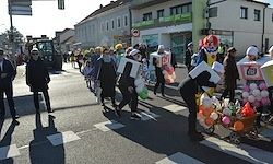 Eine Gruppe von Menschen in Kostümen geht auf der Straße, einige mit Luftballons. Eine Person beugt sich nach unten, um etwas aufzuheben. Gebäude befinden sich auf beiden Seiten der Straße.
