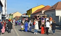 Eine Straße voller Menschen in verschiedenen farbenfrohen Kostümen. Sie gehen vorbei und versammeln sich um eine kleine Bühne oder einen Stand in der Mitte. Die Gebäude auf beiden Seiten haben verschiedene Farben und Designs.