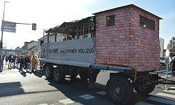 Ein LKW mit einer ziegelsteinartigen Struktur auf der Rückseite fährt auf einer Straße. Es stehen Menschen am Straßenrand. Links sind Straßenlaternen und ein Verkehrsschild.