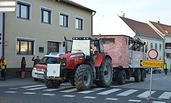 Ein roter Traktor fährt mit einem Anhänger über einen Zebrastreifen. Ein rotes Auto ist dahinter. Ein Schild steht an der Seite. Gebäude befinden sich auf beiden Seiten.
