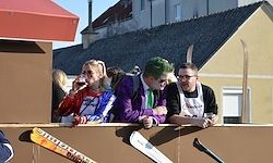 Vier Menschen, zwei Frauen und zwei Männer, in Kostümen gekleidet und mit Getränken in der Hand, stehen auf einem Balkon mit Blick auf Gebäude und Skier im Hintergrund.