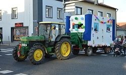 Ein grüner Traktor mit gelben Rädern fährt die Straße entlang und zieht einen dekorierten Anhänger mit Luftballons, während eine Menschenmenge zusieht.