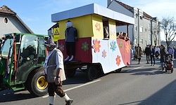 Ein Umzug mit einem geschmückten Anhänger, der von einem Traktor gezogen wird, und einem Mann in traditioneller Kleidung, der neben ihm geht, sowie mehrere Fußgänger beobachten.