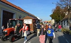 Eine Gruppe von Menschen läuft auf einer Straße mit einem roten Traktor und einem kleinen Anhänger. Einige Leute tragen Winterkleidung, und es gibt Gebäude auf beiden Seiten.