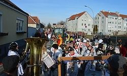 Eine Menschenmenge in Kostümen und Hüten versammelt sich um eine Brass-Tuba auf einer Straße, mit Gebäuden und einer Straßenlaterne im Hintergrund.