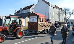 Ein Traktor zieht einen kleinen, hölzernen Paradewagen die Straße entlang, wo Menschen auf dem Bürgersteig gehen. Gebäude und Bäume säumen die Straße.