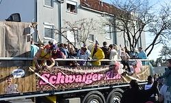 Ein Festwagen voller Menschen in gelben Kostümen, einer hält ein Banner mit der Aufschrift 'Schwager Kids', mit Zuschauern auf der Straße.