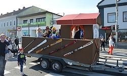 Eine Gruppe von Menschen fährt in einem Anhänger, der wie ein Haus geformt und mit Schwertern dekoriert ist. Ein Kind steht davor. Sie sind in einer Parade auf einer Stadtstraße.