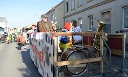 Ein Umzugswagen fährt die Straße hinunter, mit einem Mann im Anzug, einem Clown und einem Tubaspieler auf einem dekorierten Wagen. Gebäude und Menschen sind im Hintergrund.